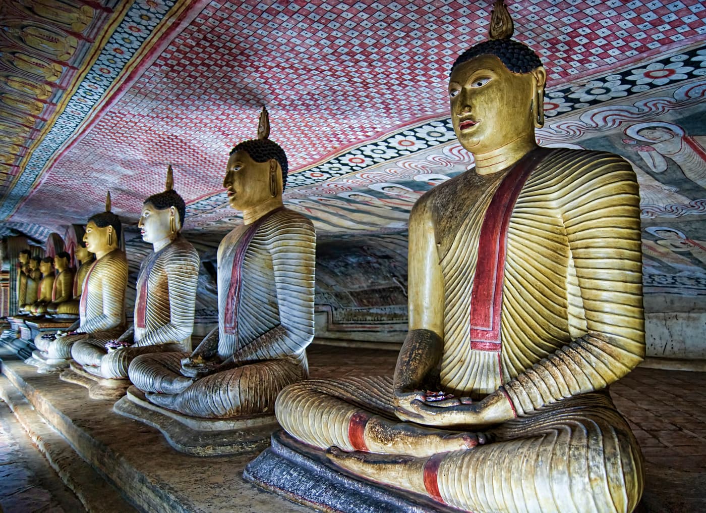 Seated golden Buddha statues in a row beneath the painted ceiling of a Dambulla cave