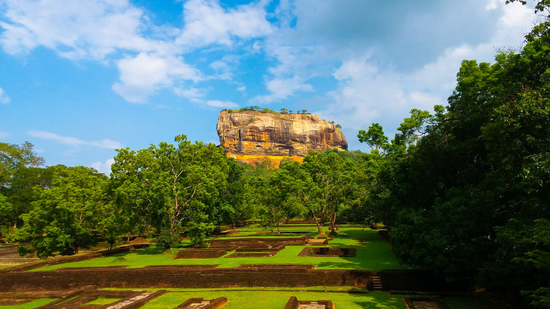 Sigiriya rock above clipped lawns and trees under a blue sky with white clouds